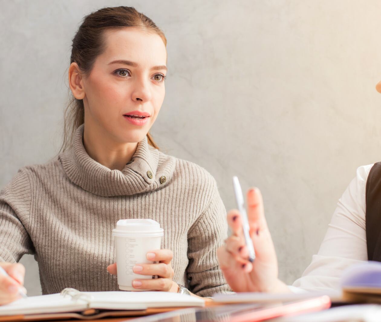 A young woman listening to her conversation partner whilst writing in a notebook and holding a takeaway coffee cup.