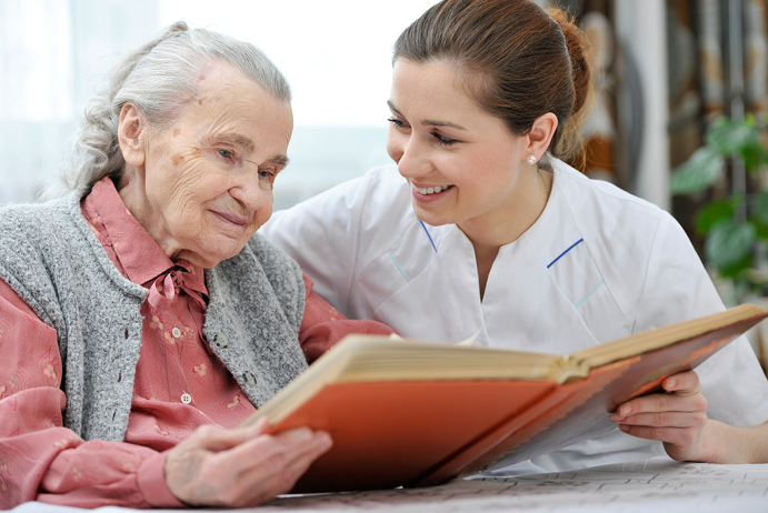 A picture of an older lady being supported by a young female healthcare assistant to read a book which they are both holding whilst smiling at the same time.