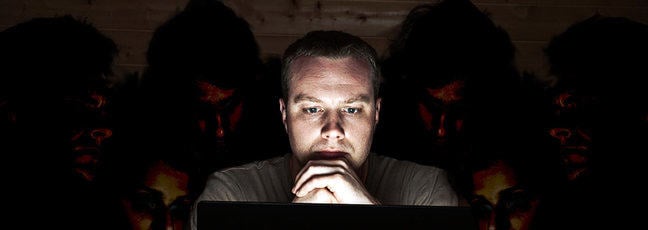 A man looks at his laptop screen, whilst others lurk behind him looking over his shoulder