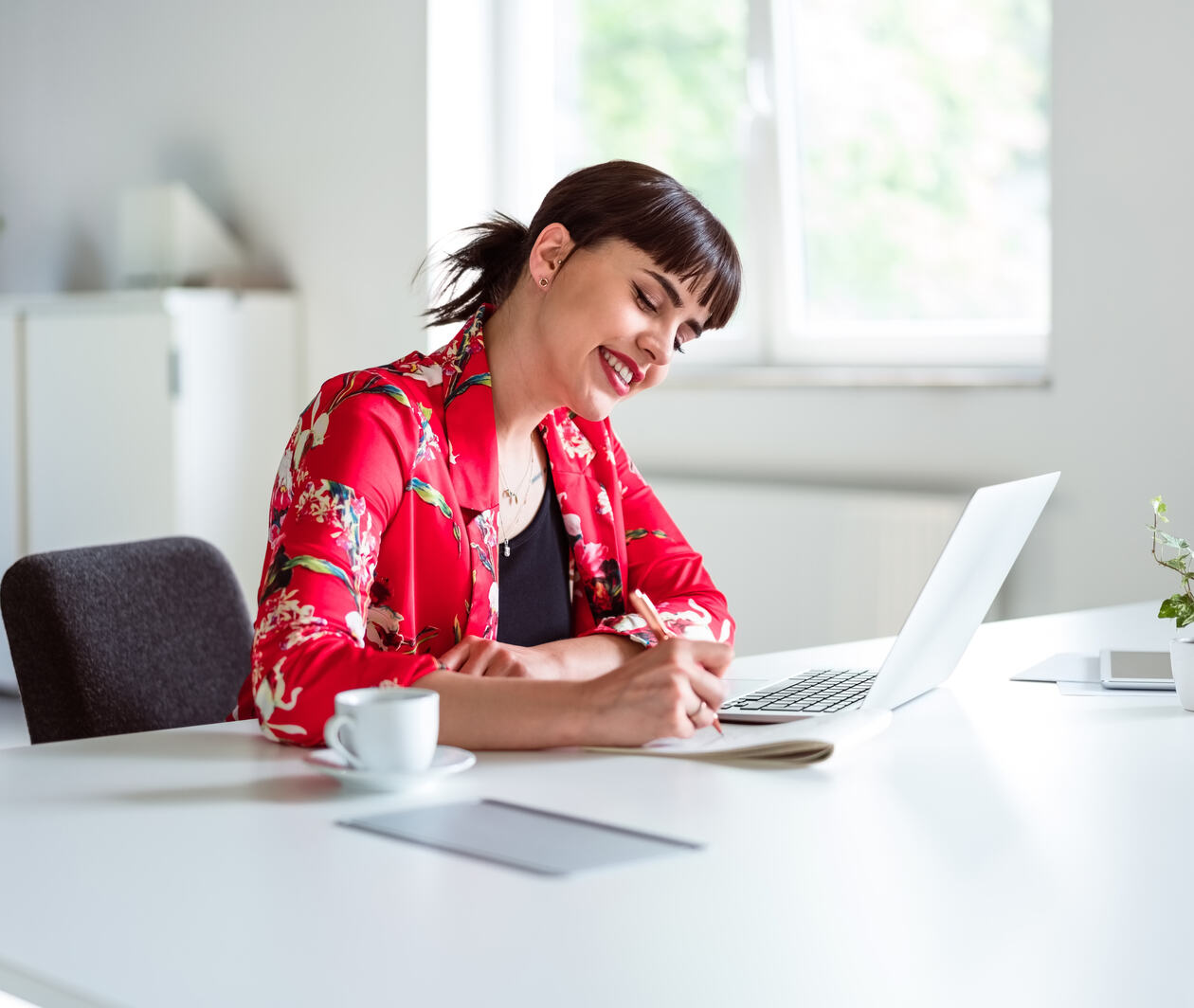 Thoughtful young woman wearing red floral jacket sitting at the desk over laptop in the creative workplace doing digital copywriting.