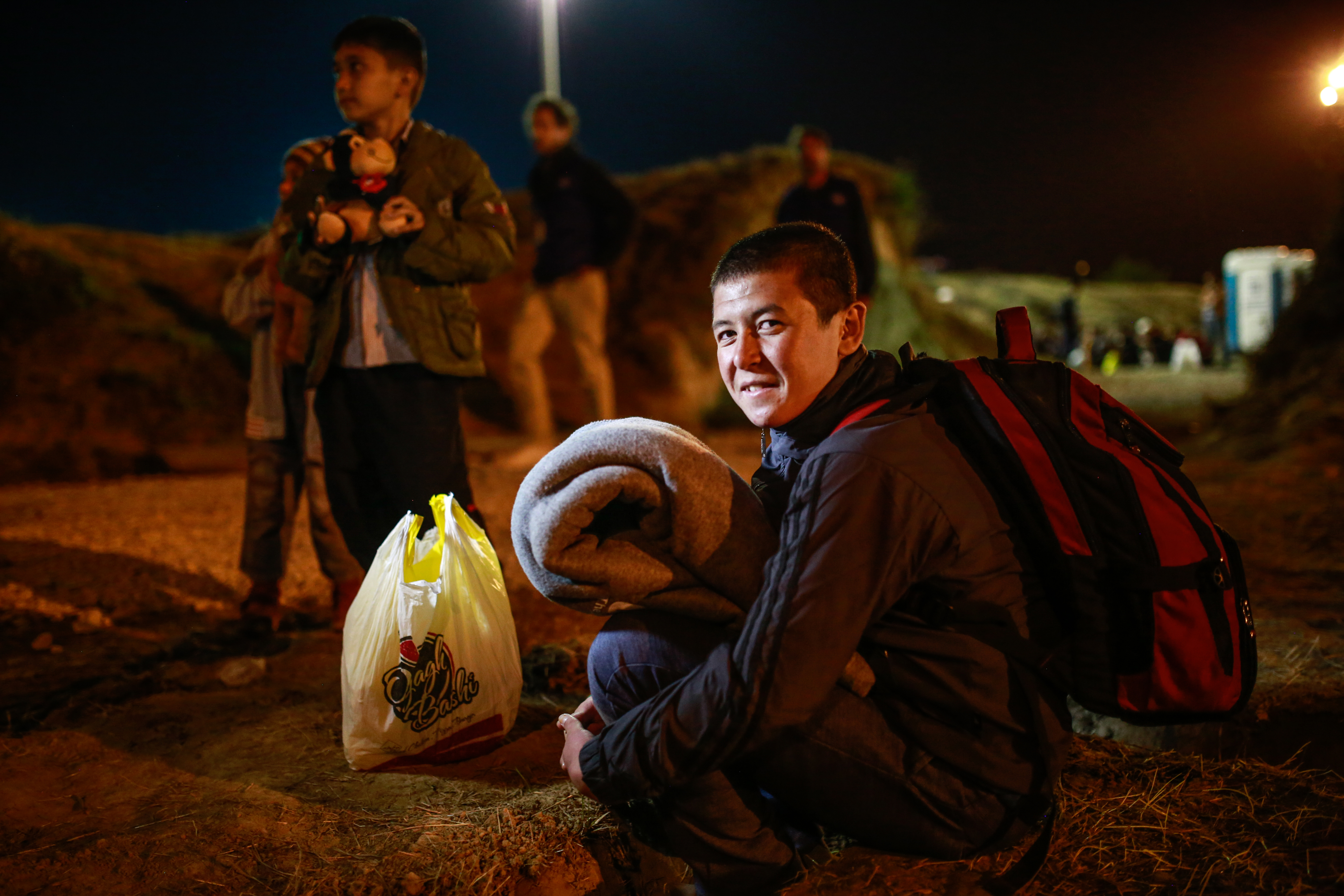 An unaccompanied teenager is crouching on the ground holding a blanket and carrying a backpack. It is night time and he is smiling at the camera. There are other unaccompanied children and adult refugees standing nearby.