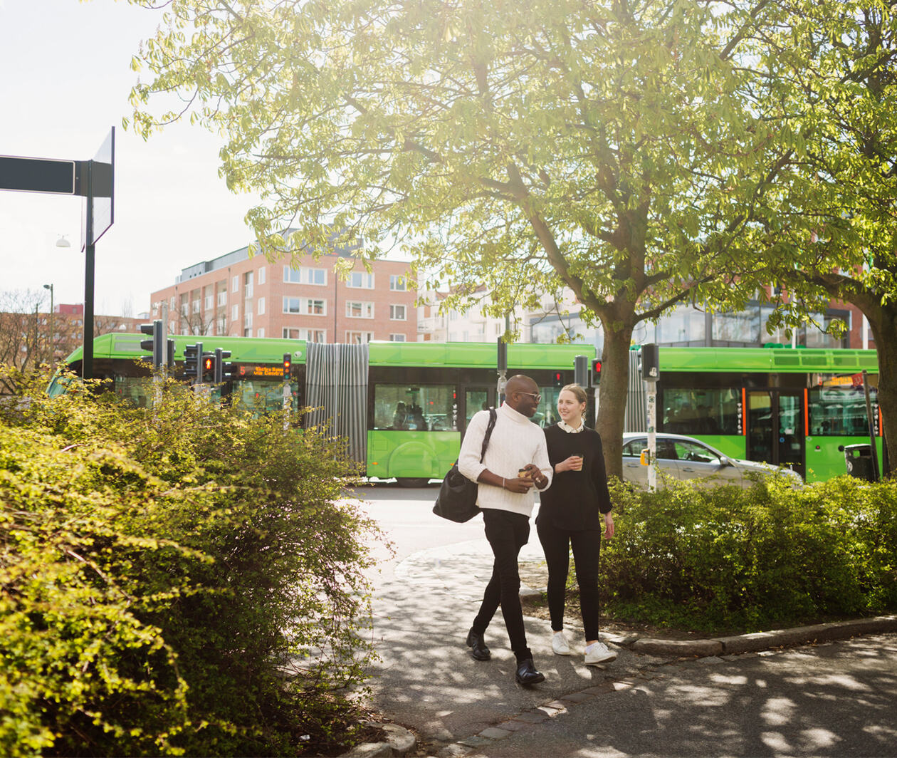 Two people walking on sidewalk against articulated bus in city, with trees and plants on the walkway, and an electric bus on the road behind.