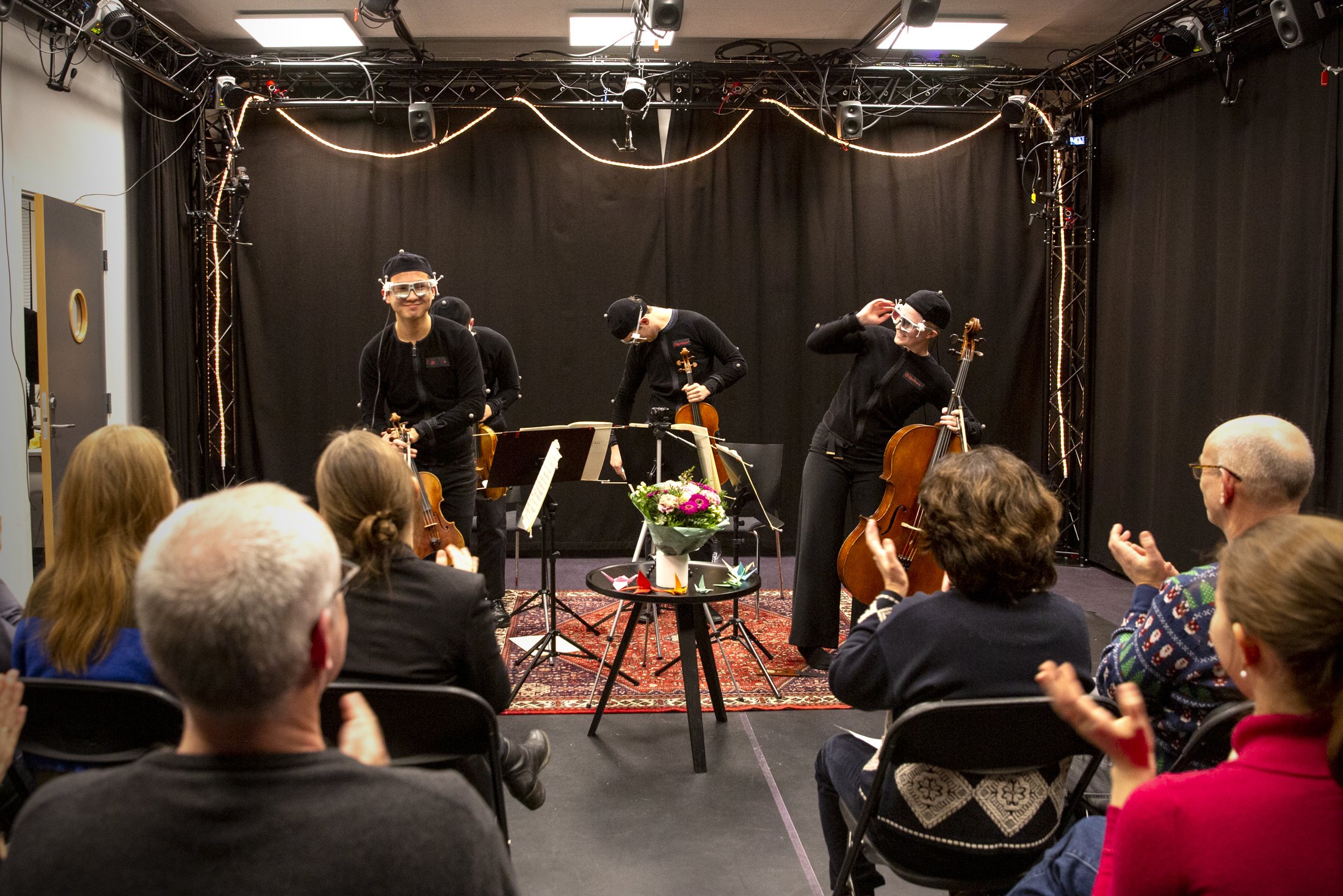 A string quartet in motion capture equipment in front of an audience. The audience is applauding.