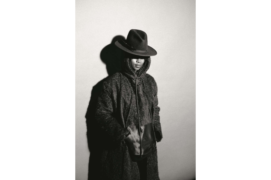 Black and white image of a woman with braids, wearing a long coat and a fedora that covers her eyes