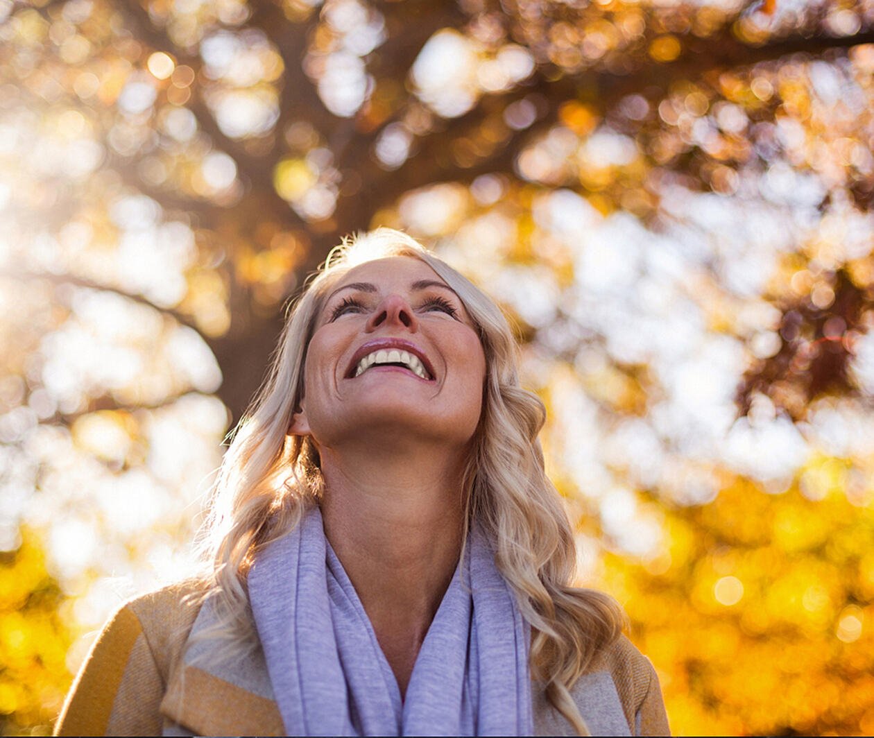 Se observa una mujer mirando al cielo y sonriendo. De fondo hay árboles y se ve el reflejo del sol.