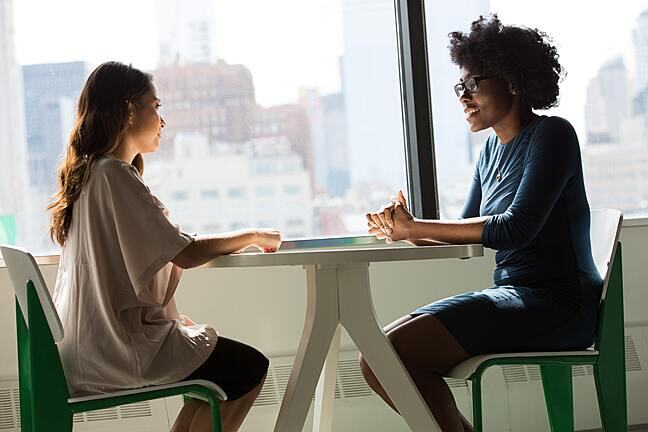 Two women sitting at a desk near a window and chatting