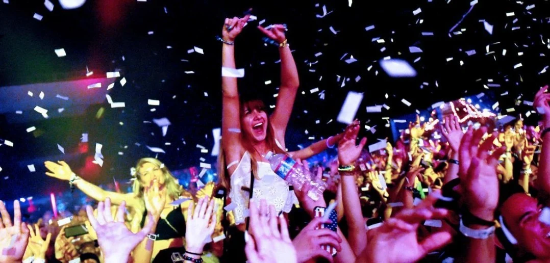 A crowd in a nightclub, covered in confetti. A woman in the centre is sitting on someone's shoulders with her arms in the air, smiling and shouting.