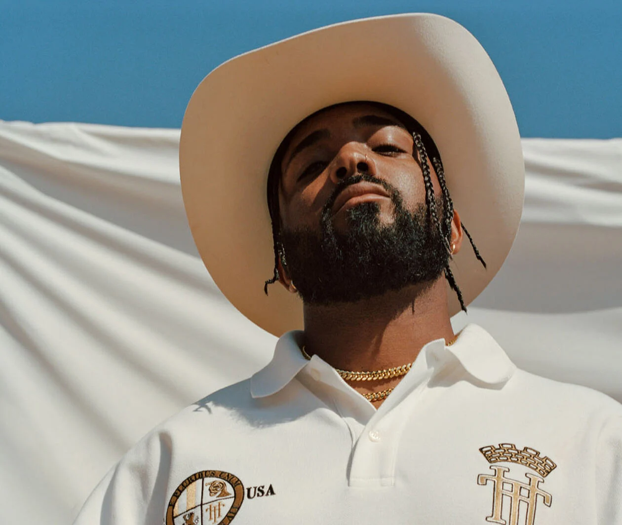 Member of the Compton Cowboys, dressed in a white shirt with a white cowboy hat standing in front of a white background with blue sky in the background