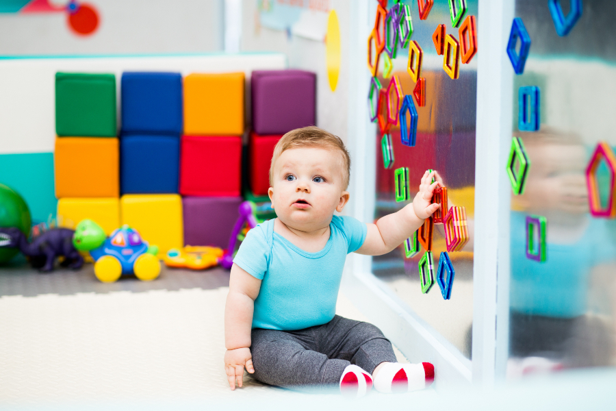 Baby boy wearing a blue top playing with magnets on the wall sitting on the floor.