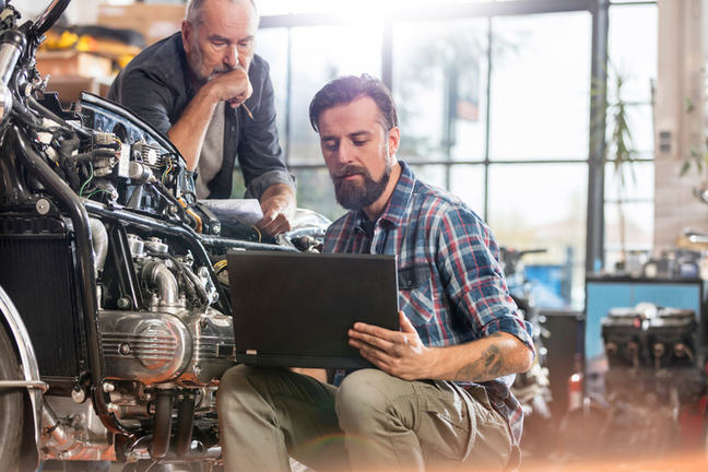 Two men looking at a laptop while working on a car engine.