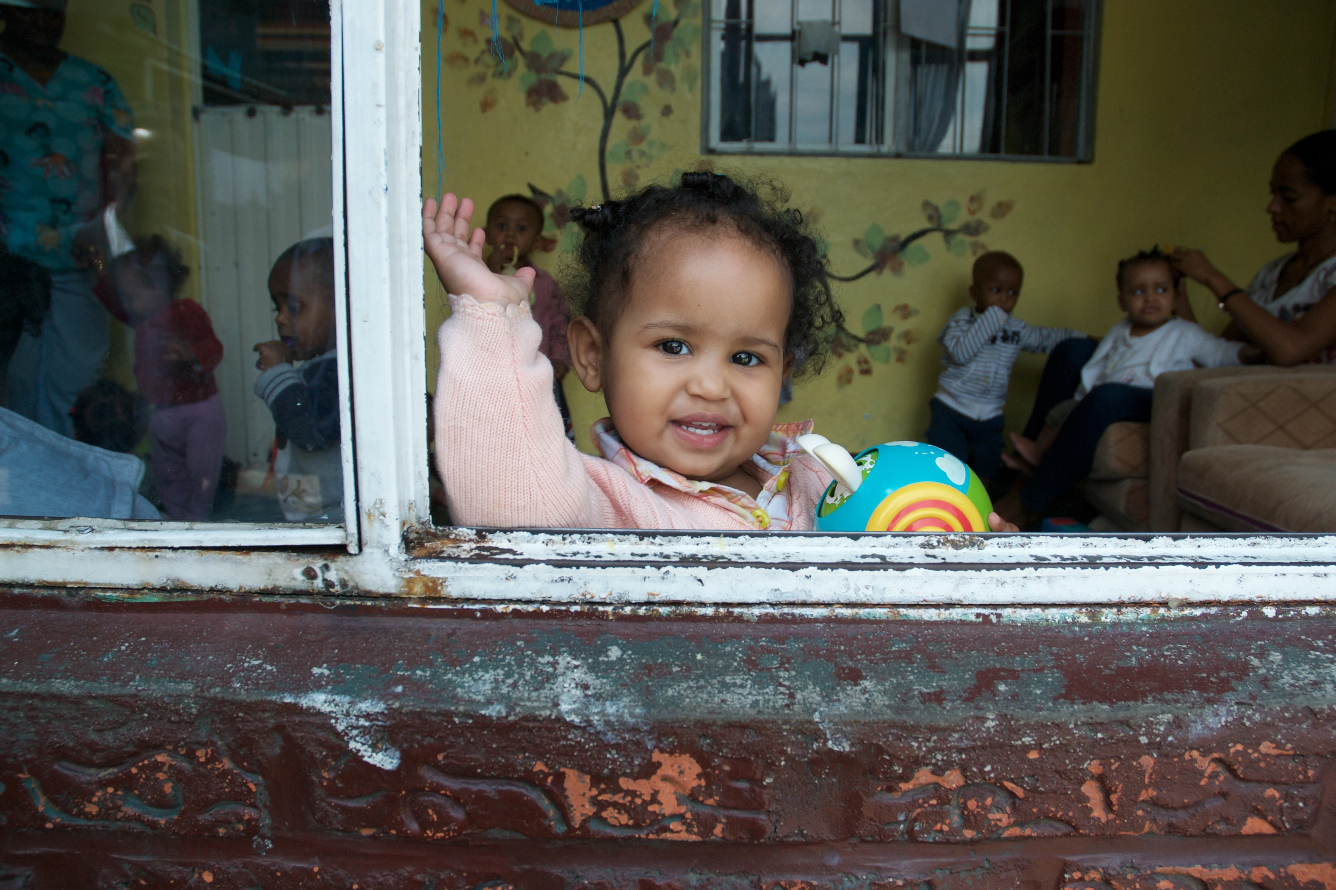 A girl leans out of the window in her daycare centre and waves to the camera.