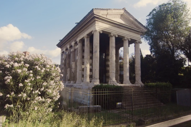 A square temple that's raised from ground level, surrounded by trees and bushes.