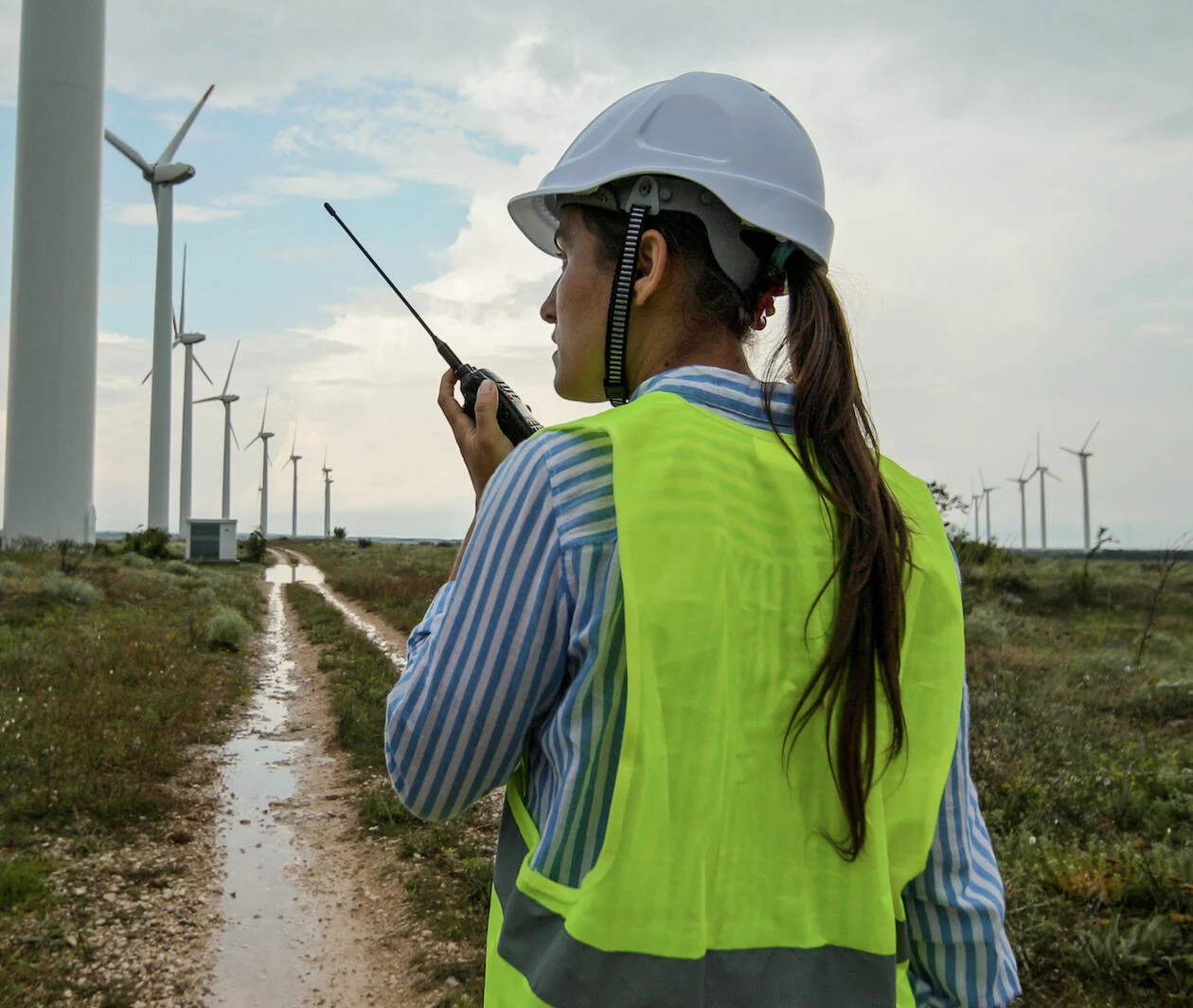 A woman in a hard hat and high-vis jacket talking into a walkie-talkie, we look over her shoulder with shots of wind turbines, open fields and a blue sky just beyond her.