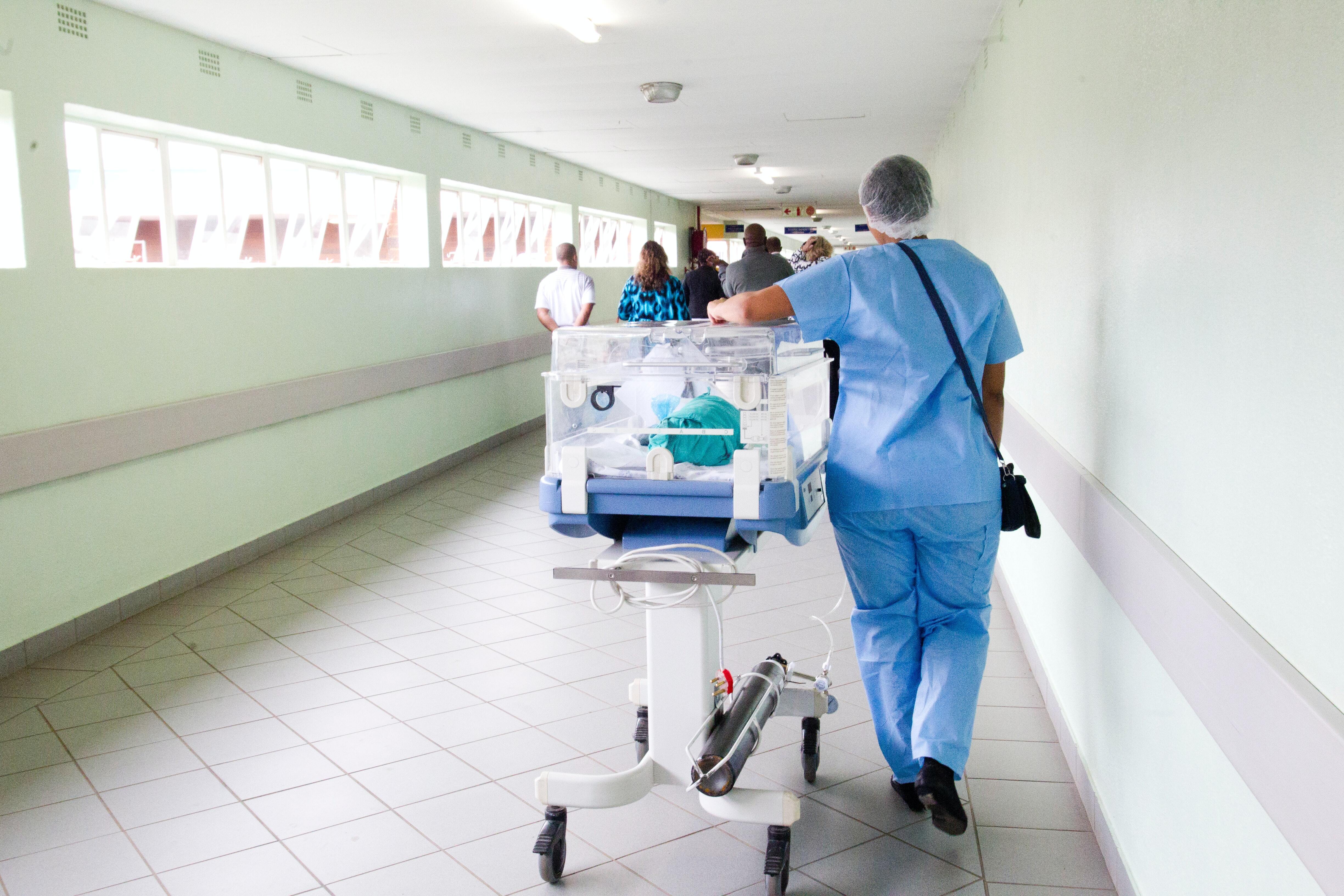 Hospital nurse wearing blue scrubs and cap shown carrying some medical equipment on a specialist table down a hospital corridor. Patients can be seen ahead, conversing with doctors at the end of the corridor, which is bright green.