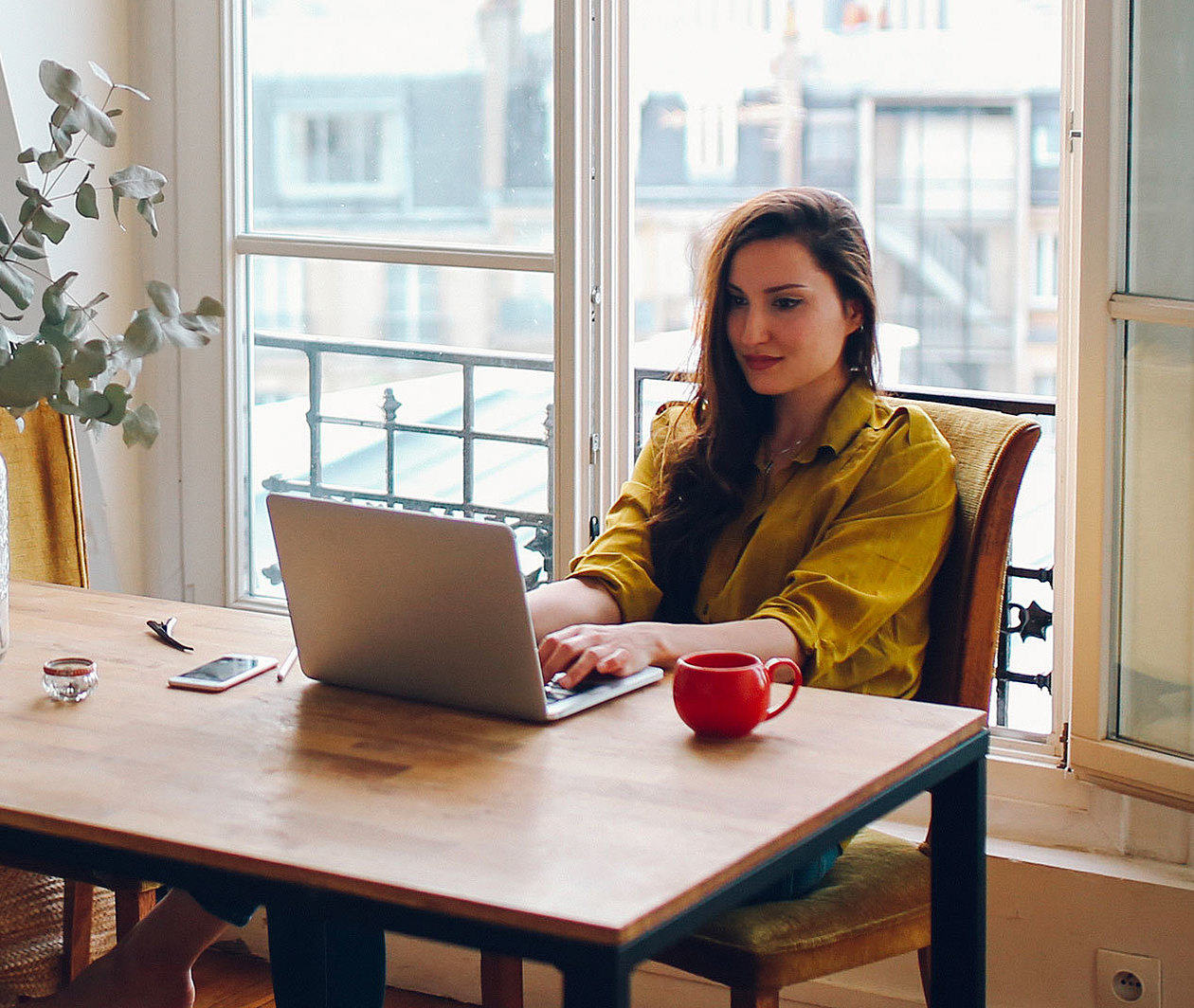 A photograph showing a person seated at a computer in frot of a window, beyond which is an open landscape featuring blurred green trees with hills in the background. The person sits with their back to us and is wearing headphones. They have bare feet.