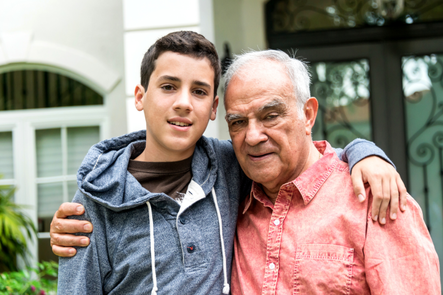 Boy in grey blue hoodie with his arm around the shoulder of older man in pink shirt, both standing in front of an older style house.