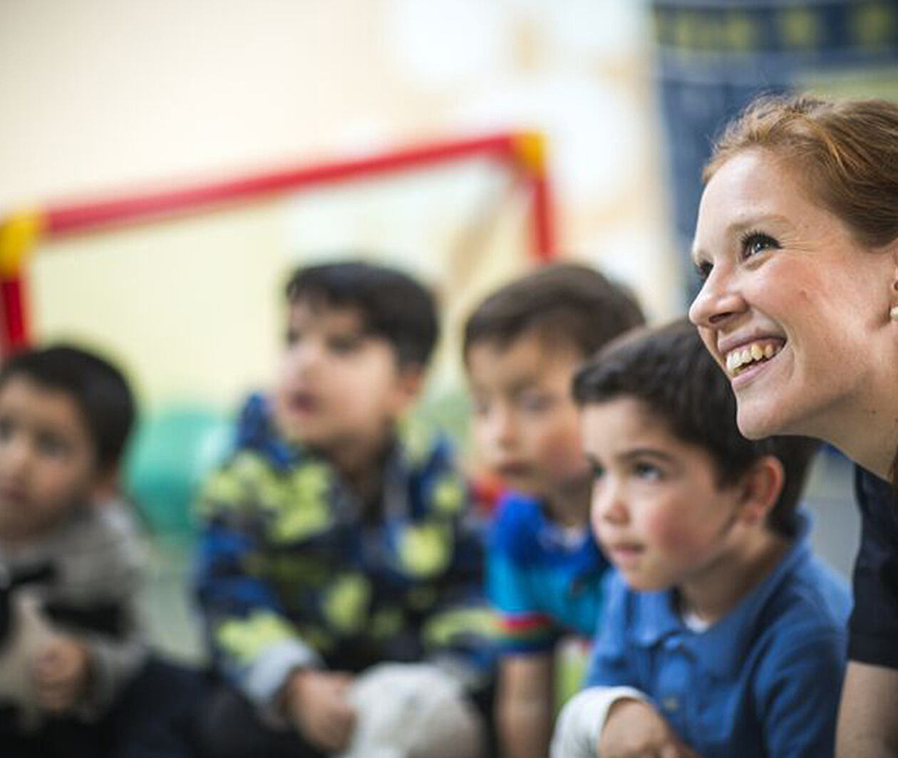 Smiling teacher sat alongside young students, all looking in the same direction at something off screen