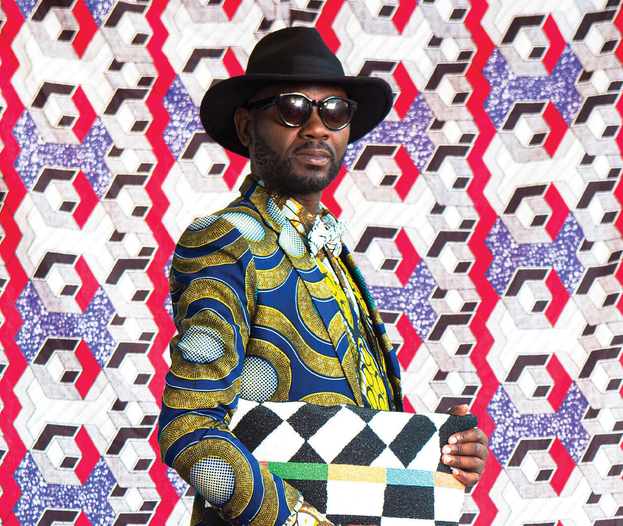 A man poses dressed in a colorfully patterned suit with a large hat, sunglasses and bag.