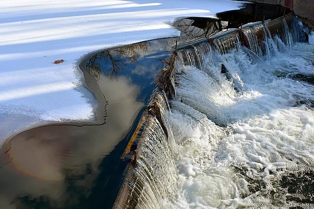 Image of water pouring over a dam from a large, ice-covered river.