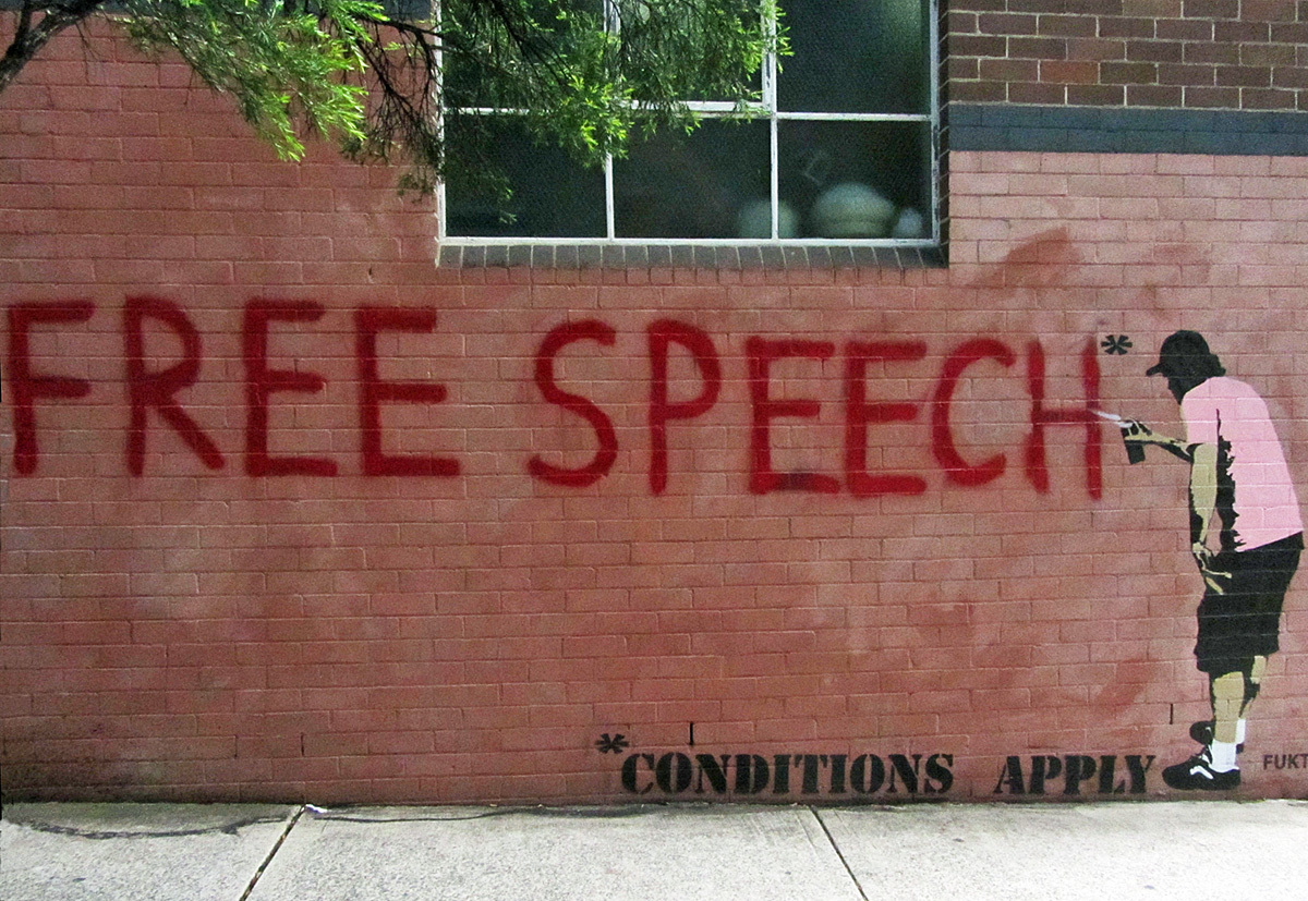 Banksy graffiti of a man spray painting 'Free Speech *conditions apply' on a brick wall.