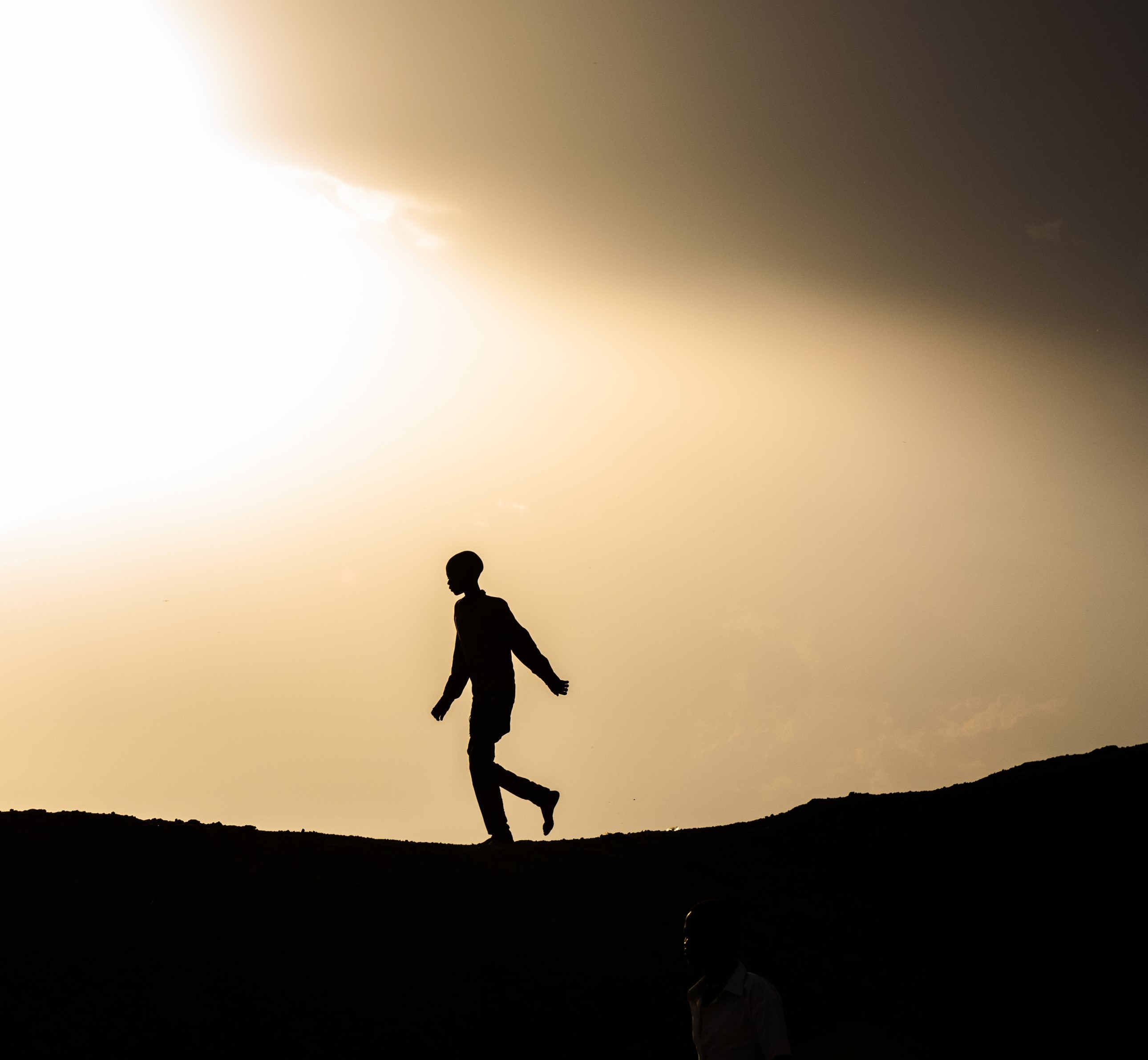 A silhouette of a boy with a long shirt and long pants walking on top of a hill.