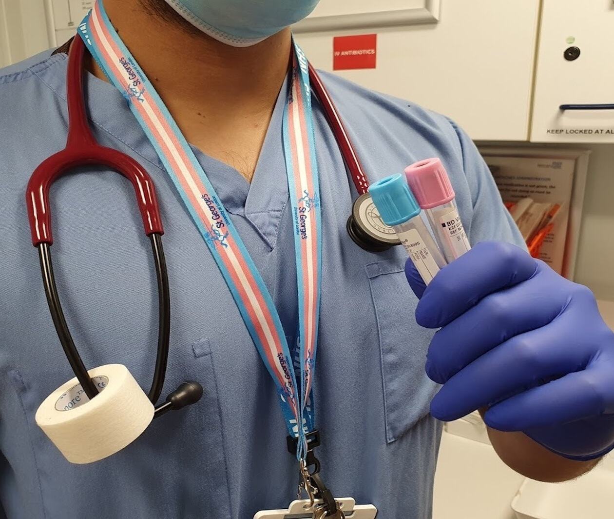 A medical professional wearing blue scrubs, stethoscope with a white tape roll on it, a transgender flag colour lanyard and two medical tubes with the trans blue and pink colour tops on them   