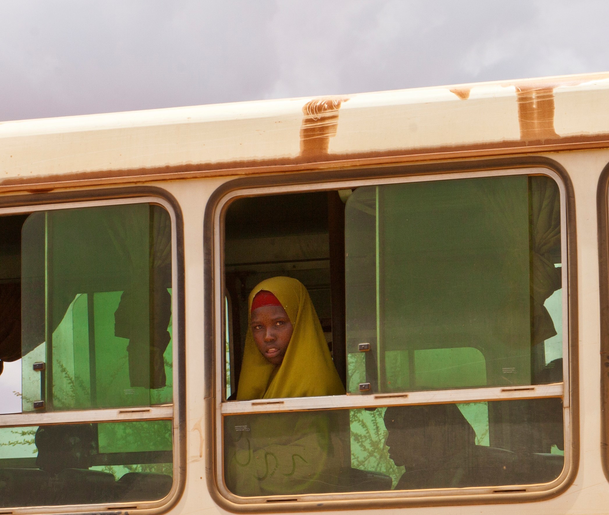 A young African girl with yellow material covering her head and shoulders is standing in a bus looking out of the window. There are silhouettes of other children in the bus sitting nearby.