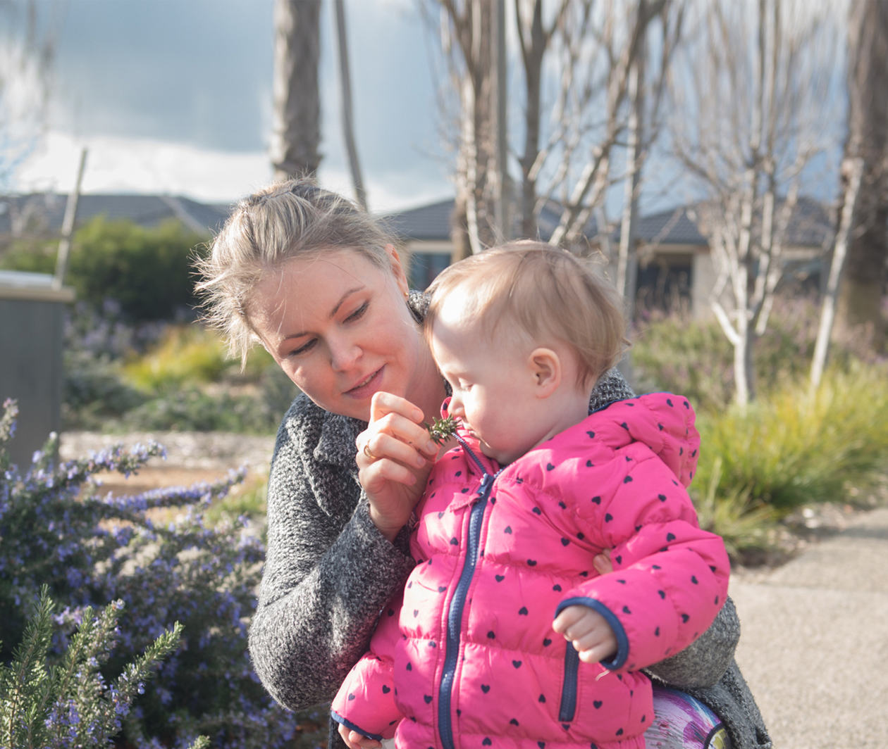 The photo is of a female cancer patient walking on a garden path holding her young infant daughter. In the photo, the patient is holding a flower up to her daughter's nose. 