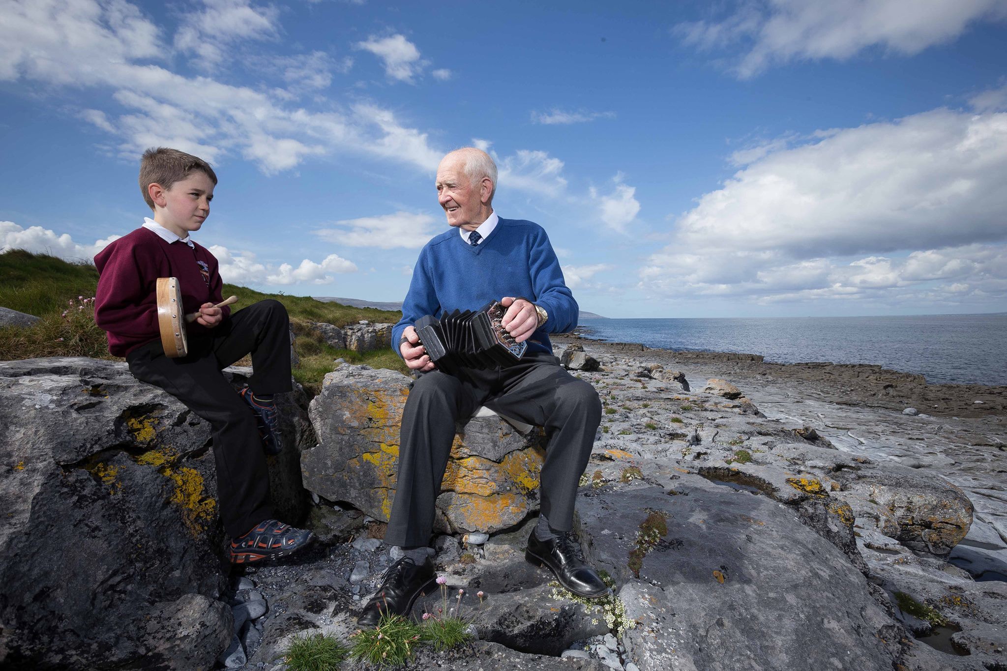 A man and a young boy sitting on rocks on a beach playing musical instruments