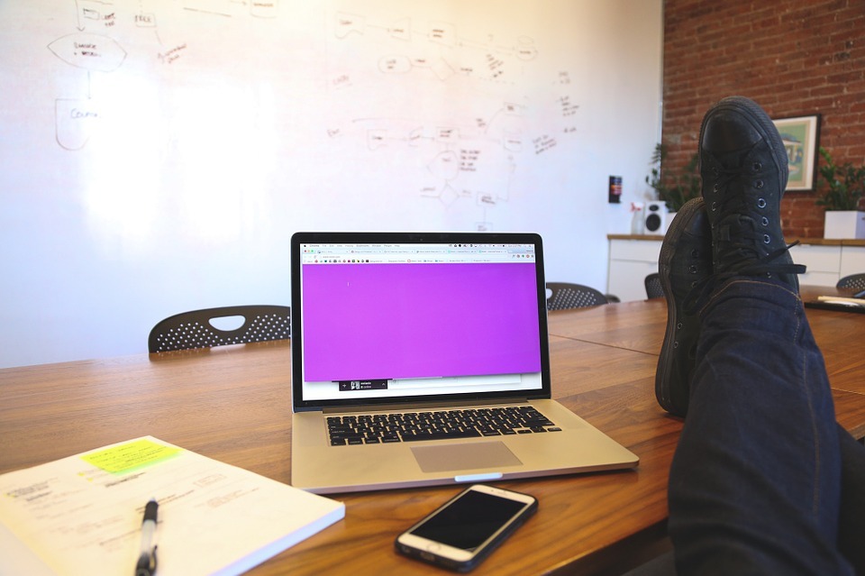 A man's feet on a desk, with a laptop, phone and notebook against the background of a whiteboard with lots of diagrams.