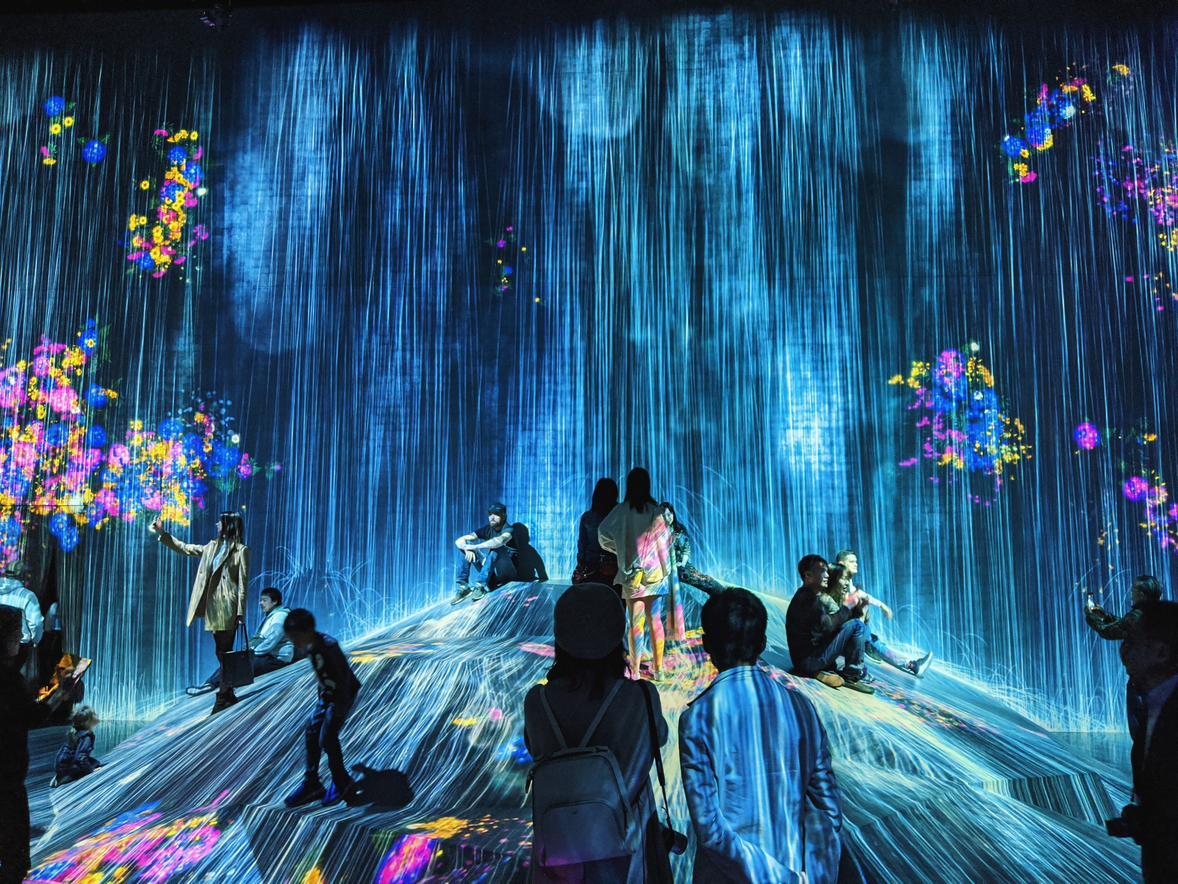 Inside an art exhibition a group of people are sitting and standing on and around a mound. Falling rain and coloured arrays of flowers are projected onto the whole scene including the people.