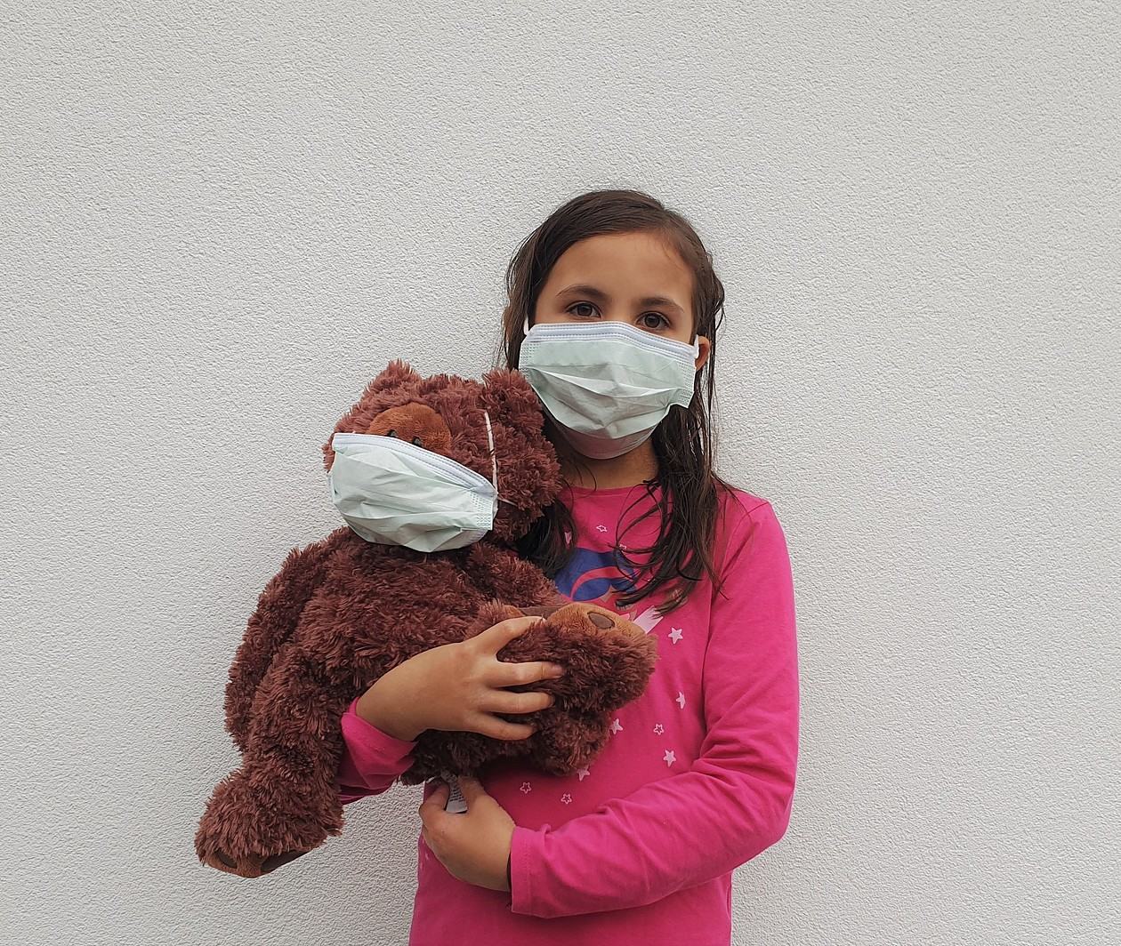 Young girl holding a brown teddy. Both are wearing a face mask.