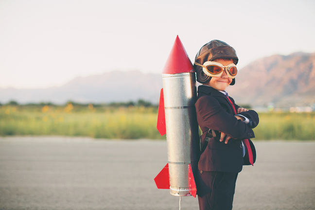 A young business-minded boy is wearing a business suit, space helmet with a rocket strapped to his back. His hands are on his hips and he is smiling at the camera standing on blacktop.