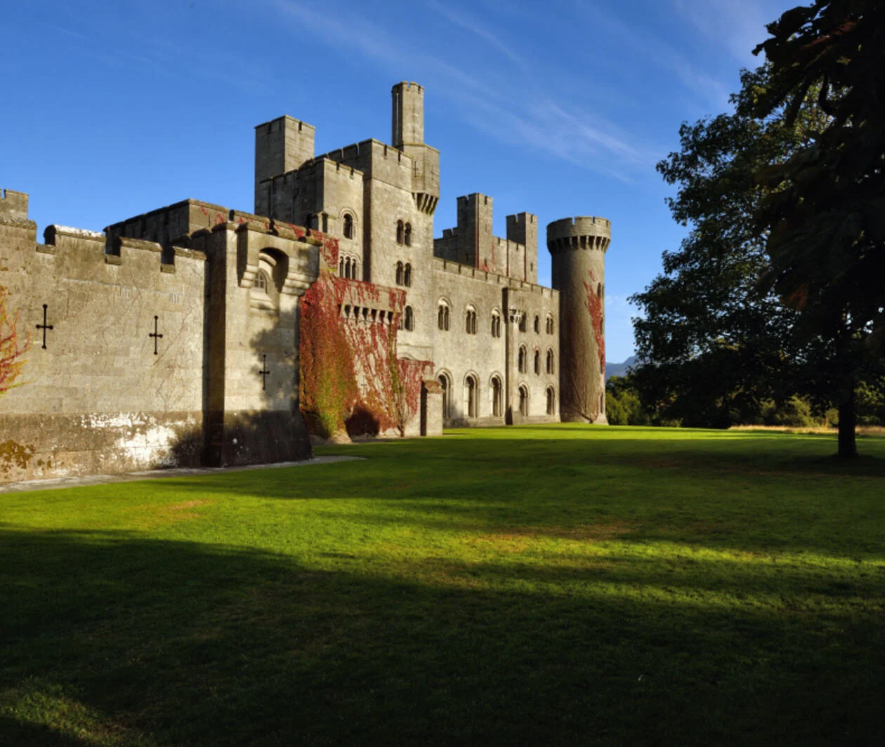 Penrhyn Castle on a sunny day with green grass and trees in front and a bright blue sky.