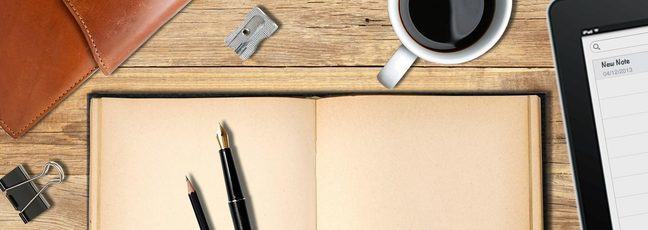 A pen and pencil sit on an open notebook with a coffee and an iPad close at hand, ready to start writing fiction