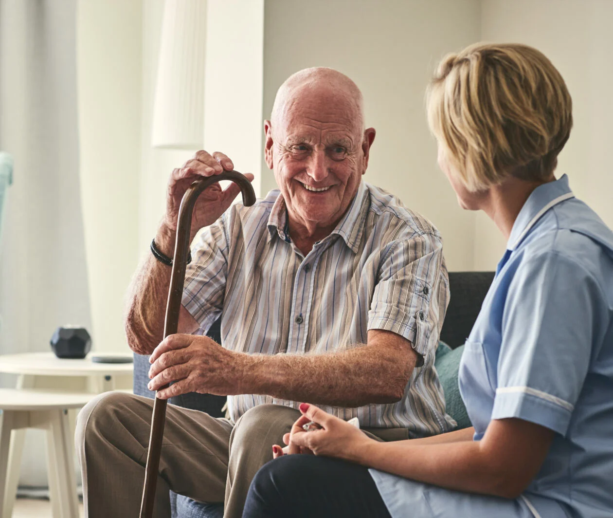 Elderly man with a walking stick, sat talking to a nurse in a care home. 