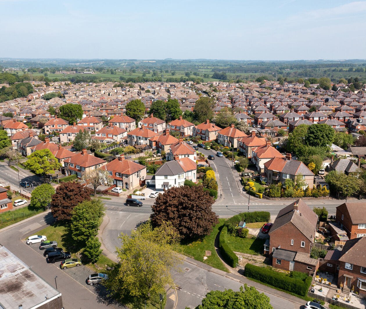 A view from above of a suburb: Roads, houses, a farm, green fields and a community centre.