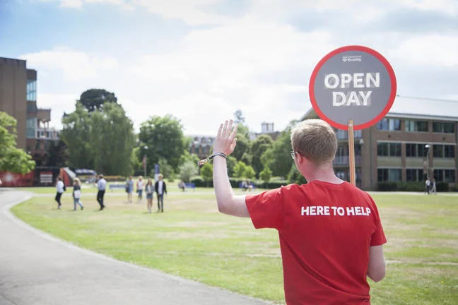 A student holding a sign saying Open Day and wearing a t-shirt with the writing here to help is ready to help prospective students during their visit.