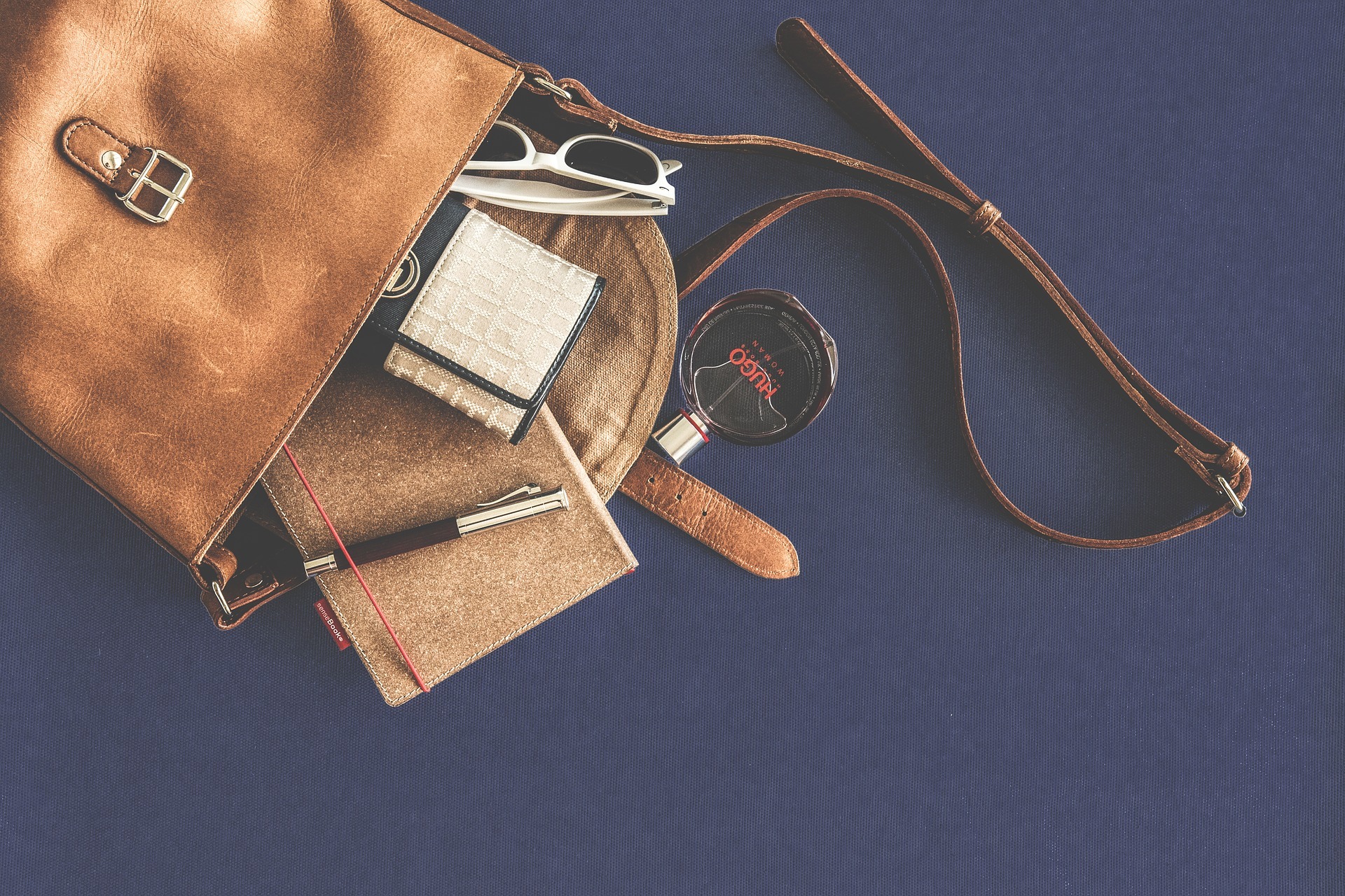 A bag with items spilling out of it - sunglasses, wallet, pen and notebook, perfume