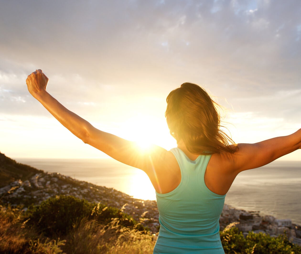 Woman standing on a hill at sunrise with arms raised in celebration, overlooking the ocean and a coastal town below.