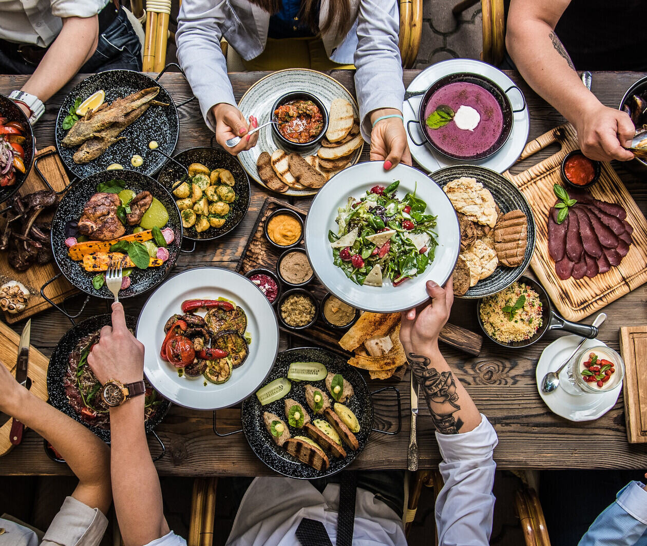 A group sit at a large, dark brown table with dozens of plates of food. They help themselves.