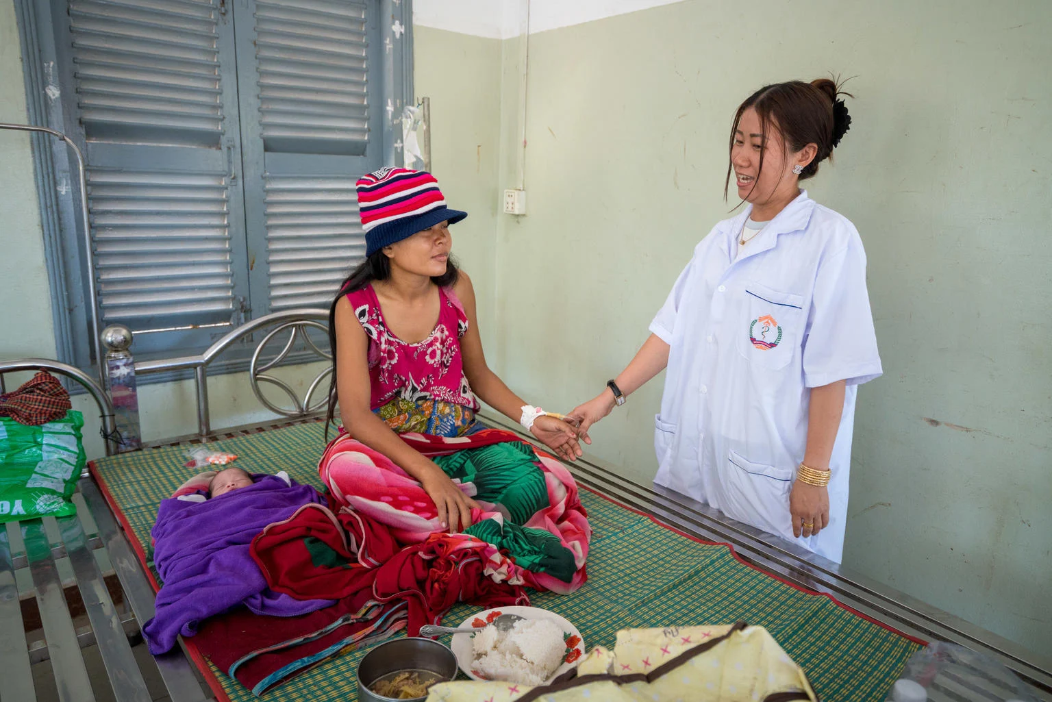 A female nurse holds the hand of a female patient sitting on a bed.