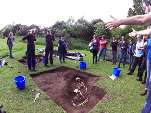 A forensic anthropologist explains a grave site with a body partially excavated to a group of students