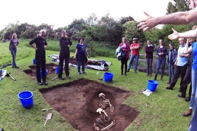 A forensic anthropologist explains a grave site with a body partially excavated to a group of students