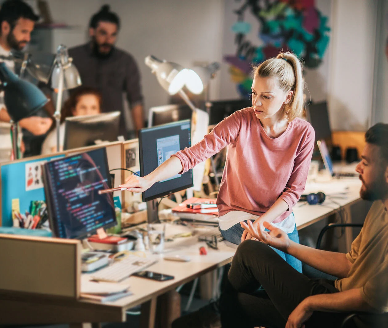 Group of computer programmers working in the office. Focus is on blond woman showing something to her colleague on PC relating to code.