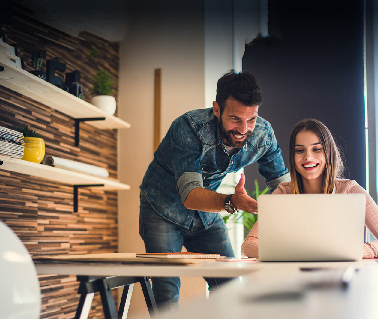 A man and a woman sit behind a laptop, smiling.