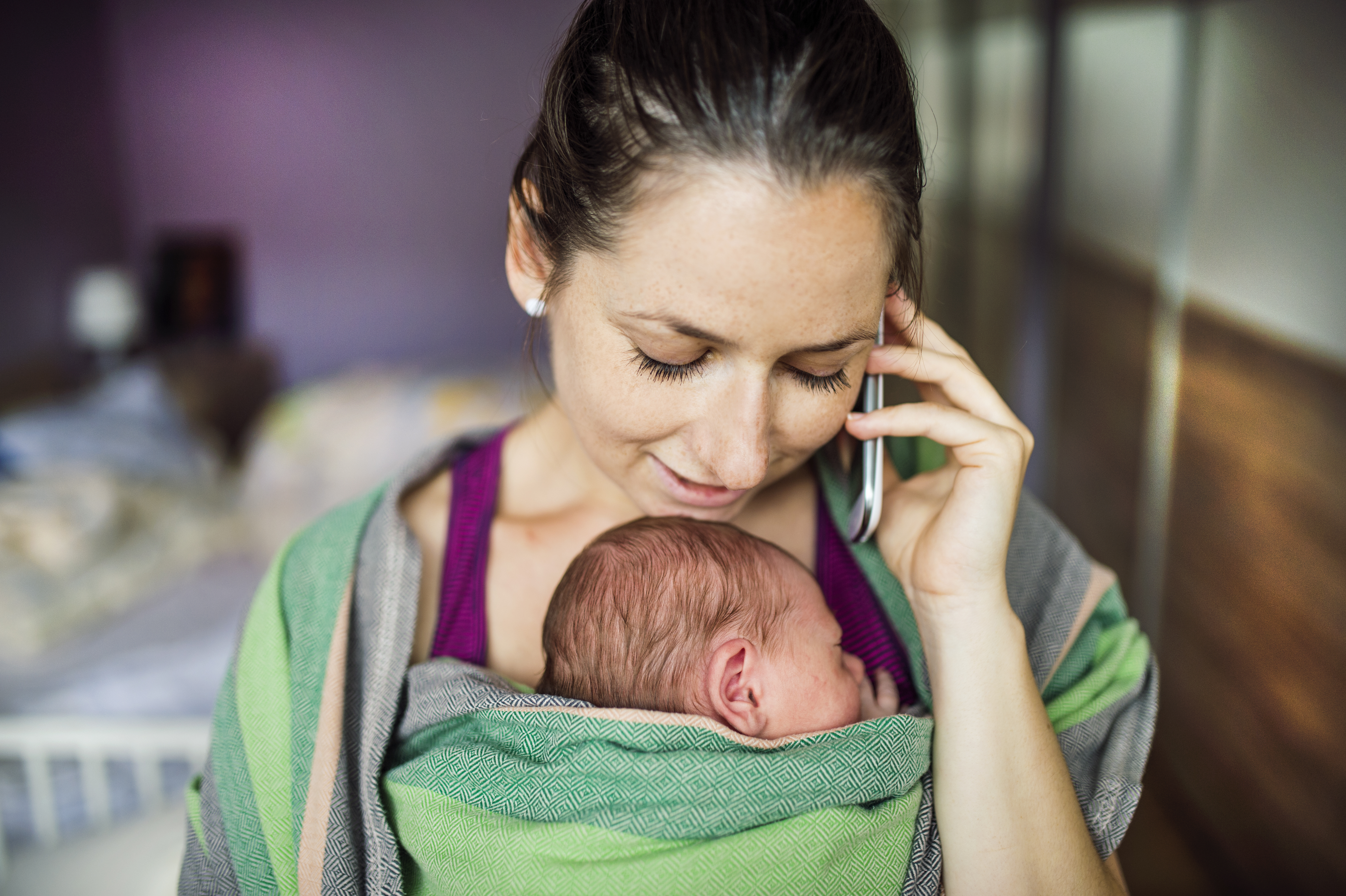 A woman with her baby in a baby carrier making a phone call with her mobile phone.