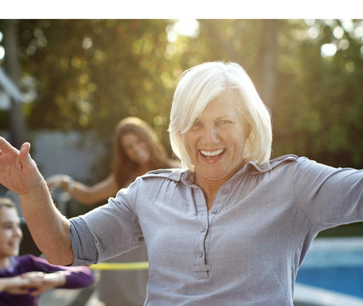 A playful image of an older woman, a teenager and young girl having fun hula hooping in a back garden next to a swimming pool.