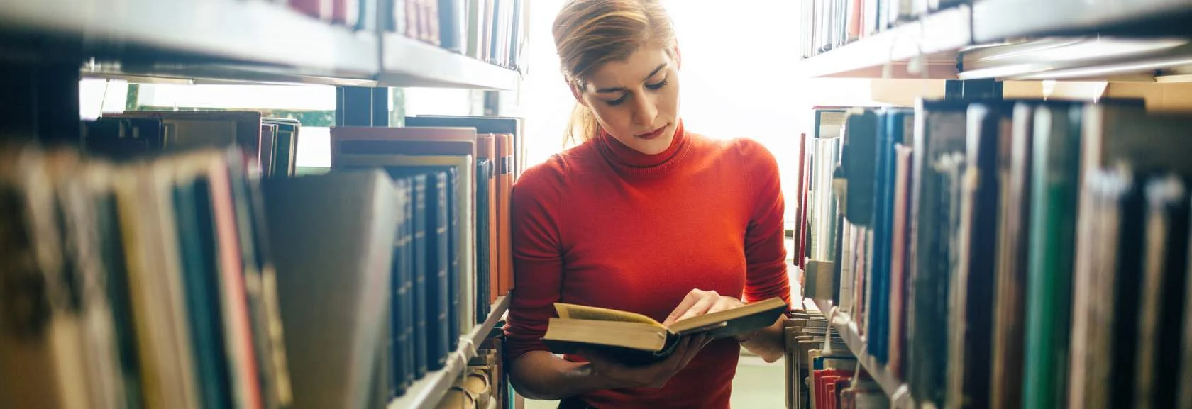 Young woman reads a book between library shelves