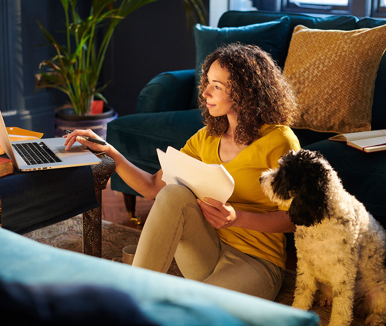 A woman sat on the floor next to her dog on her laptop. 